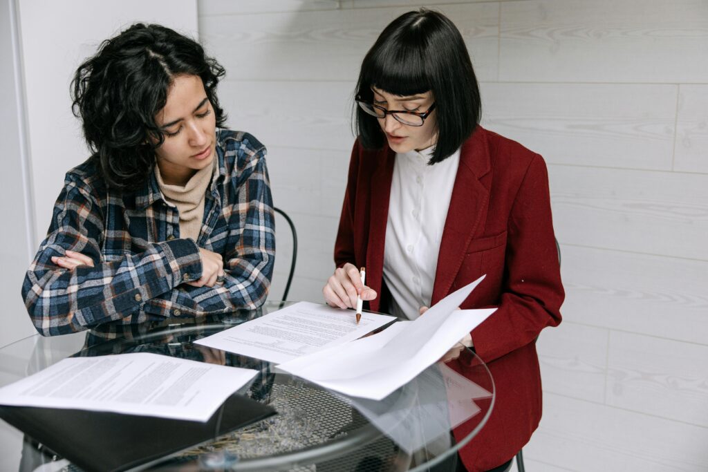 Two women collaborating at a glass table, reviewing business documents in a modern office setting.